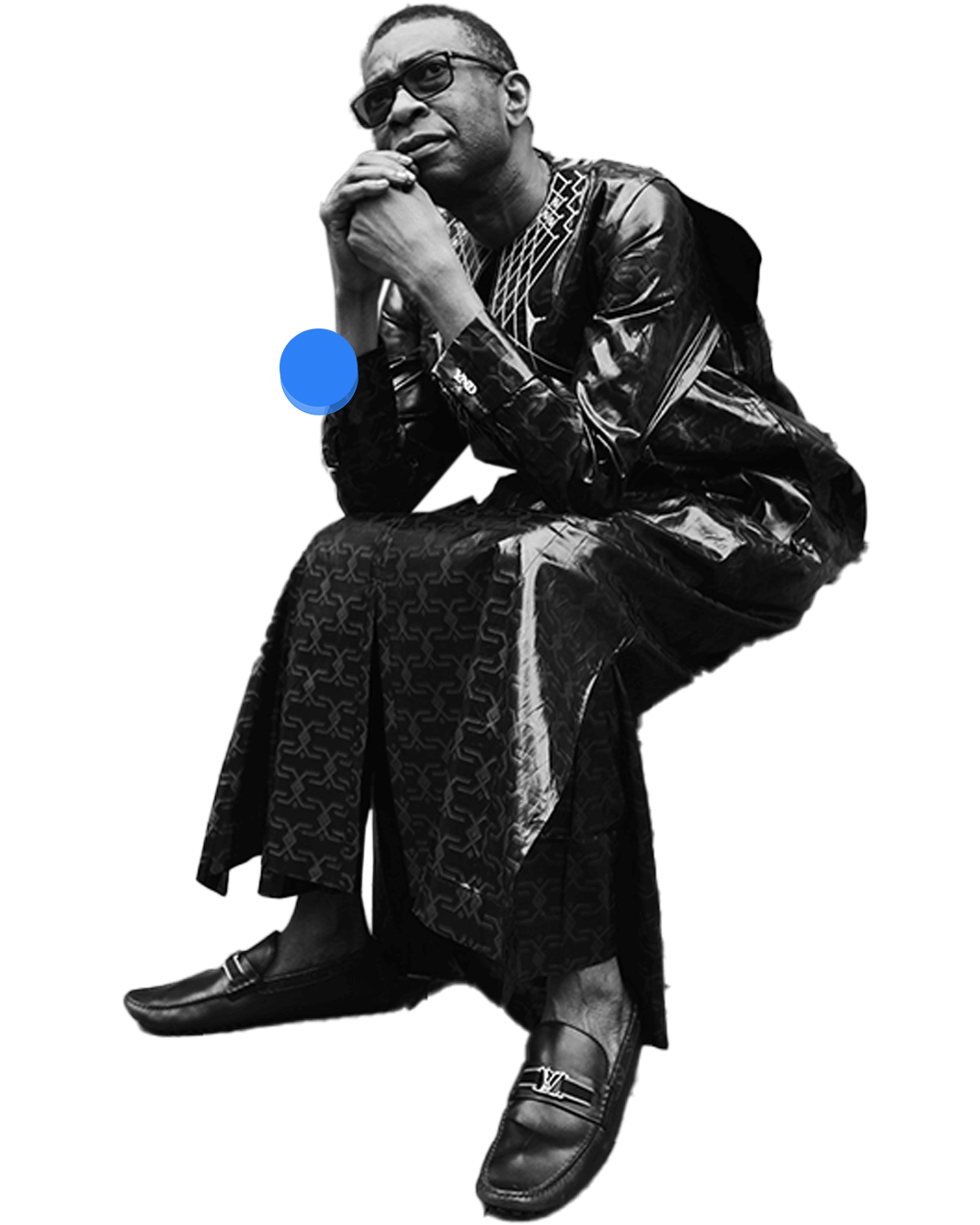 Man seated and leaning forward with hands clasped, wearing patterned traditional attire and glasses.